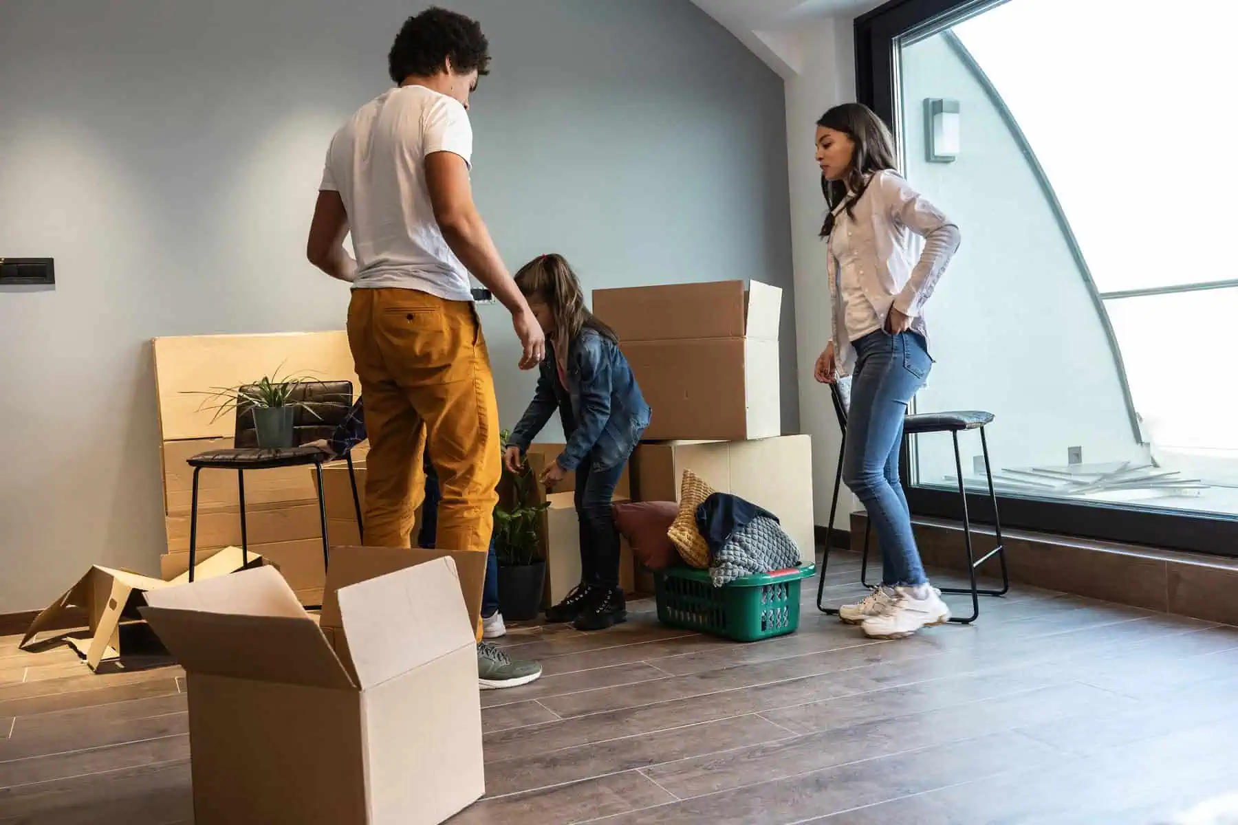 Stokarea - Mini Armazens Self storage Em uma sala com piso de madeira e janelas grandes, três pessoas estão ocupadas desempacotando. Elas estão arrumando plantas e vários itens de seu aluguel de self-storage em Stokarea enquanto caixas de papelão estão espalhadas, transformando o espaço em um refúgio de organização. Lidar em Portugal