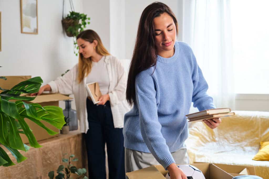 Stokarea - Mini Armazens Self storage Duas mulheres estão desempacotando caixas em uma sala de estar iluminada, que elas organizaram recentemente após usar o self-storage Stokarea. Uma mulher de suéter azul segura livros, enquanto a outra de camisa branca coloca itens em uma prateleira. O cômodo exala aconchego com plantas e luz natural fluindo. Lidar em Portugal